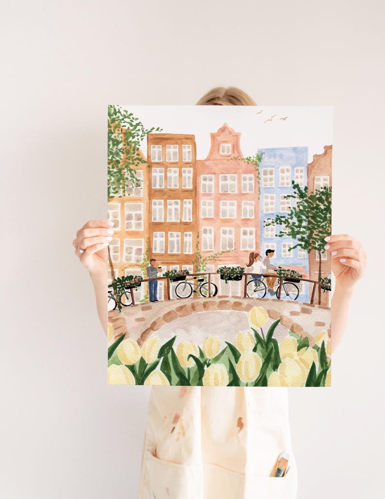 May include: A watercolour art print depicting a canal scene with tall, narrow buildings in shades of brown, peach, and blue. A stone bridge features people on bicycles and a person standing, with yellow tulips in the foreground and green trees.