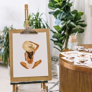Op de afbeelding: Een aquarel schilderij van een persoon die een grote strohoed en een witte badpak draagt, zittend met gekruiste benen. Het schilderij staat op een ezel met een houten frame.