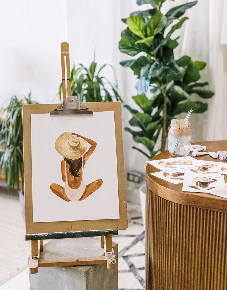 Op de afbeelding: Een aquarel schilderij van een persoon die een grote strohoed en een witte badpak draagt, zittend met gekruiste benen. Het schilderij staat op een ezel met een houten frame.