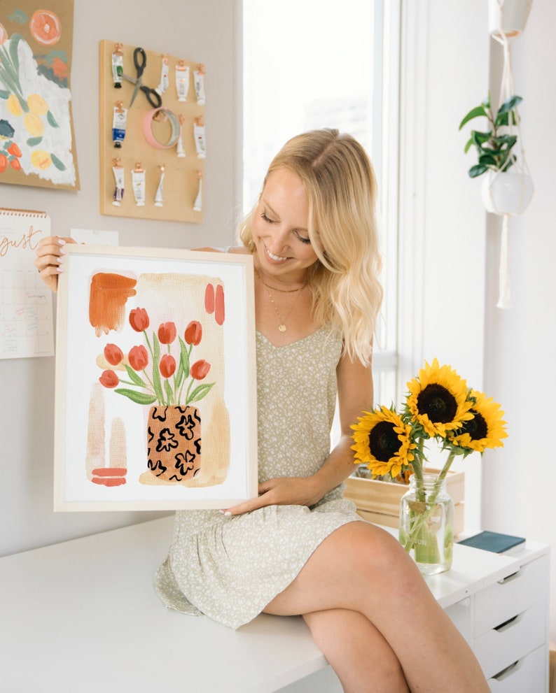 Puede incluir: Una mujer sostiene una pintura acuarela enmarcada de tulipes rojas en un jarr&oacute;n con un patr&oacute;n negro y marr&oacute;n. La pintura tiene un fondo marr&oacute;n claro con formas abstractas en naranja y rojo.