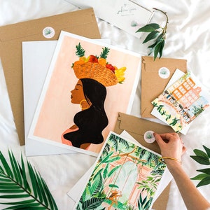 Puede incluir: Un conjunto de tres impresiones de arte que representan a una mujer con una cesta de fruta en la cabeza, una escena de jard&iacute;n tropical y una escena de calle de la ciudad. Las impresiones est&aacute;n en papel blanco y tienen un fondo rosa claro.