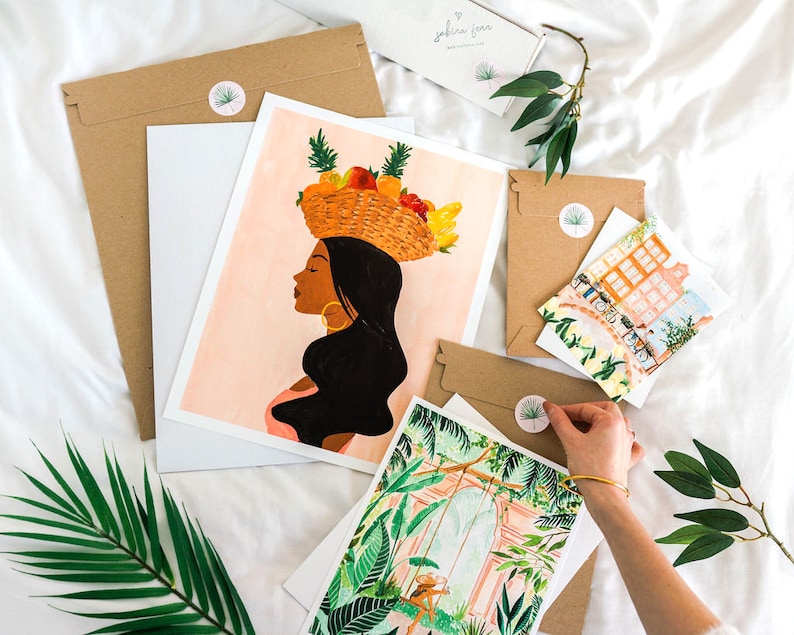 Puede incluir: Un conjunto de tres impresiones de arte que representan a una mujer con una cesta de fruta en la cabeza, una escena de jard&iacute;n tropical y una escena de calle de la ciudad. Las impresiones est&aacute;n en papel blanco y tienen un fondo rosa claro.