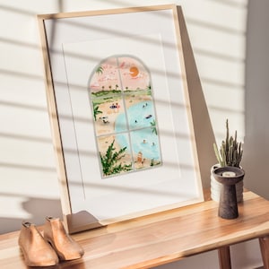 May include: A framed watercolor painting of a beach scene viewed through an arched window. The painting depicts a sandy beach with people swimming and sunbathing, palm trees, and a bright orange sun in the sky.