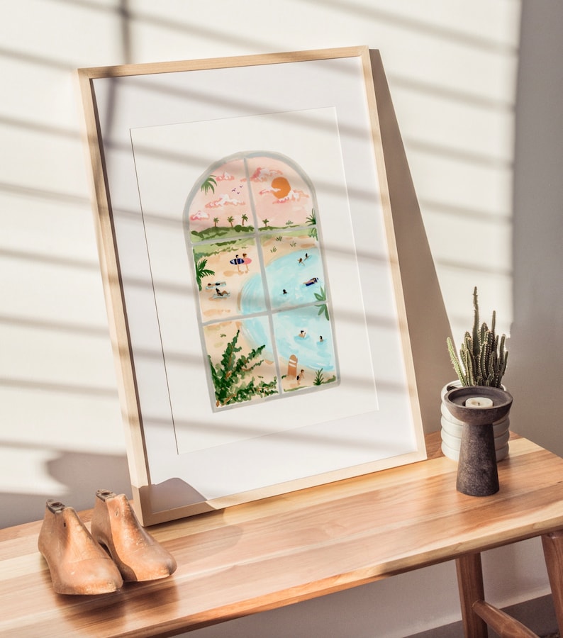 May include: A framed watercolor painting of a beach scene viewed through an arched window. The painting depicts a sandy beach with people swimming and sunbathing, palm trees, and a bright orange sun in the sky.