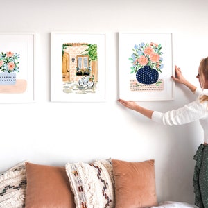 Puede incluir: Tres acuarelas enmarcadas colgadas en una pared blanca. Las pinturas representan arreglos florales en jarrones, un edificio con una puerta y una ventana, y una bicicleta.