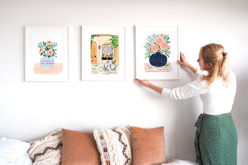 Puede incluir: Tres acuarelas enmarcadas colgadas en una pared blanca. Las pinturas representan arreglos florales en jarrones, un edificio con una puerta y una ventana, y una bicicleta.