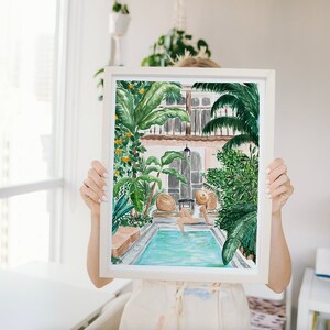 K&ouml;nnte beinhalten: Ein Aquarellbild einer Frau, die sich in einem Pool entspannt, umgeben von &uuml;ppigem Gr&uuml;n. Der Pool ist von einer Terrasse mit ein paar St&uuml;hlen und einem Tisch umgeben. Die Frau tr&auml;gt einen wei&szlig;en Badeanzug und einen Strohhut. Das Bild ist in einem wei&szlig;en Rahmen gerahmt.