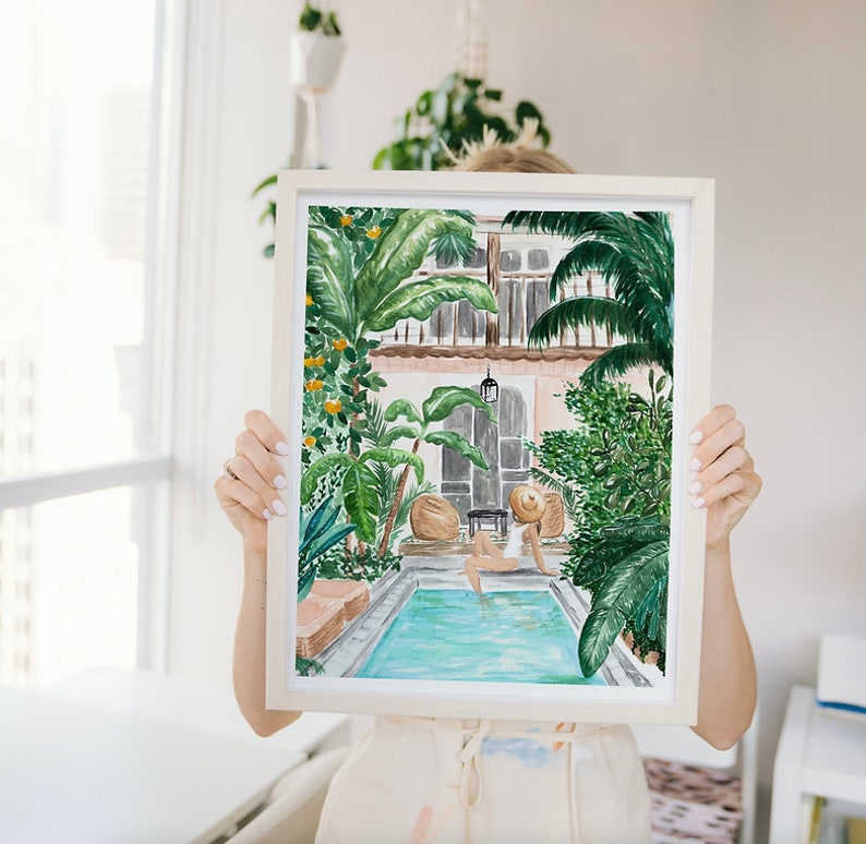 K&ouml;nnte beinhalten: Ein Aquarellbild einer Frau, die sich in einem Pool entspannt, umgeben von &uuml;ppigem Gr&uuml;n. Der Pool ist von einer Terrasse mit ein paar St&uuml;hlen und einem Tisch umgeben. Die Frau tr&auml;gt einen wei&szlig;en Badeanzug und einen Strohhut. Das Bild ist in einem wei&szlig;en Rahmen gerahmt.