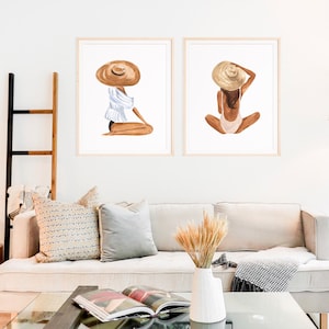 Op de afbeelding: Twee ingelijste aquarel illustraties van vrouwen die strohoeden dragen. De vrouw links draagt een wit-blauw gestreept shirt en zit met gekruiste benen. De vrouw rechts draagt een roze badpak en zit met gekruiste benen.