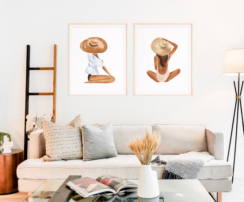 Op de afbeelding: Twee ingelijste aquarel illustraties van vrouwen die strohoeden dragen. De vrouw links draagt een wit-blauw gestreept shirt en zit met gekruiste benen. De vrouw rechts draagt een roze badpak en zit met gekruiste benen.