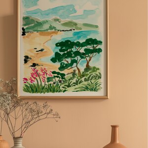 May include: A watercolor painting of a beach scene with a green tree in the foreground, blue water, and a sandy beach. The painting is framed in a gold frame and hanging on a light orange wall.