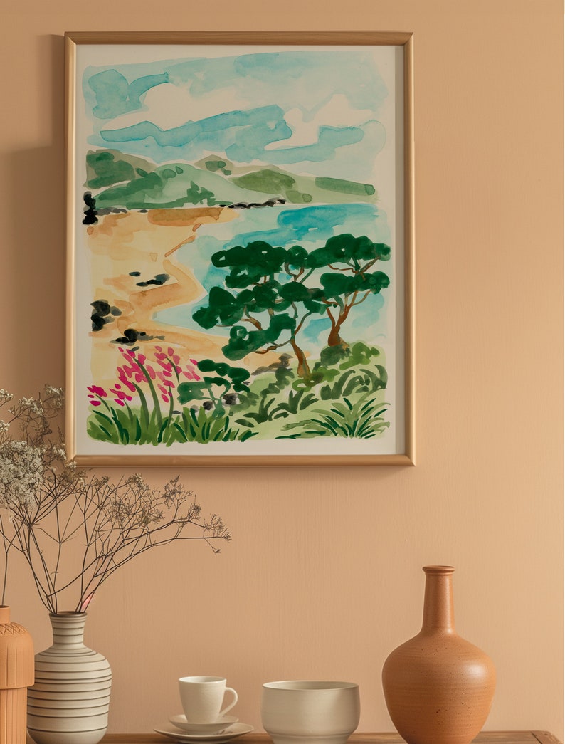 May include: A watercolor painting of a beach scene with a green tree in the foreground, blue water, and a sandy beach. The painting is framed in a gold frame and hanging on a light orange wall.