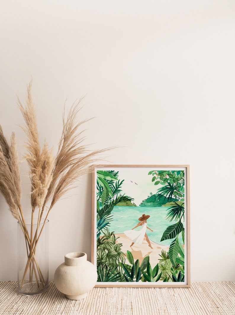 May include: A framed watercolor painting of a woman in a white dress and a brown hat walking on a beach with lush green tropical foliage in the foreground and background.