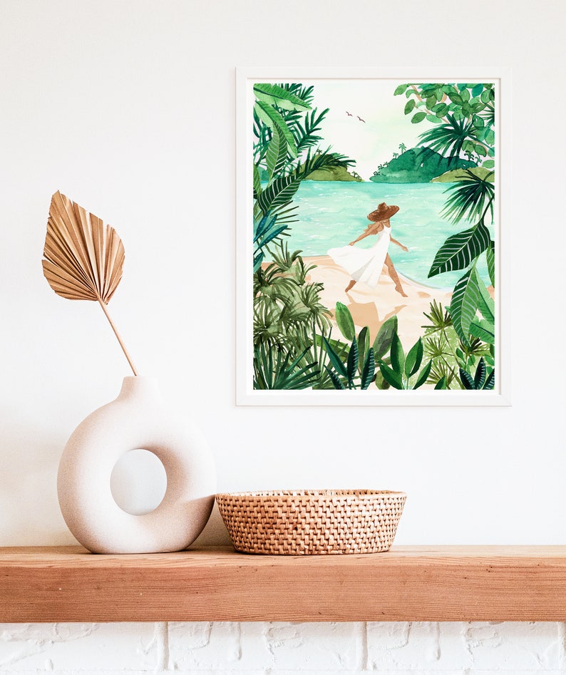 May include: A framed watercolor painting of a woman in a white dress and a brown hat walking on a beach with lush green tropical foliage on either side.