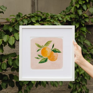 May include: A white framed print of a painting of two oranges with green leaves on a pink background.
