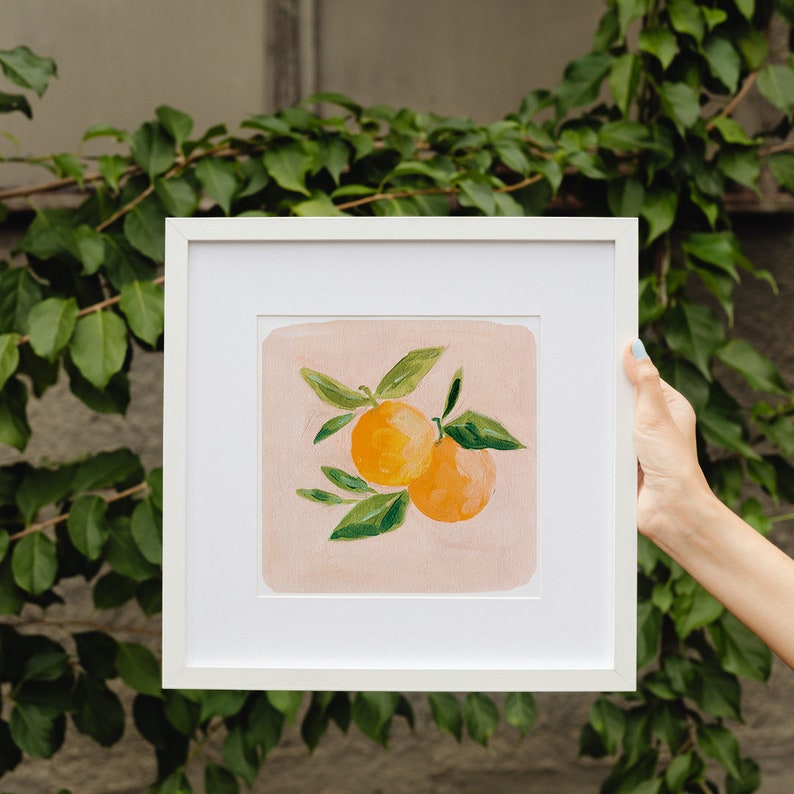 May include: A white framed print of a painting of two oranges with green leaves on a pink background.