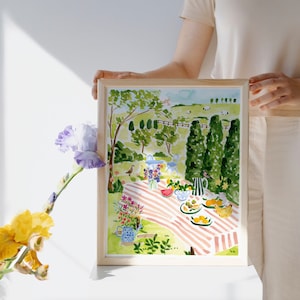May include: A watercolor painting of a picnic scene in a green meadow with a red and white striped blanket, fruit, and flowers. The painting is framed in a light wood frame.