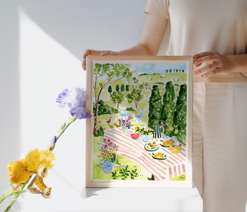 May include: A watercolor painting of a picnic scene in a green meadow with a red and white striped blanket, fruit, and flowers. The painting is framed in a light wood frame.