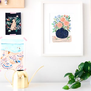 Puede incluir: Una ilustraci&oacute;n enmarcada de un ramo de flores rosas y azules en un jarr&oacute;n azul y blanco con lunares. El jarr&oacute;n est&aacute; sobre una alfombra de rayas rosas y blancas. La ilustraci&oacute;n est&aacute; enmarcada en blanco.