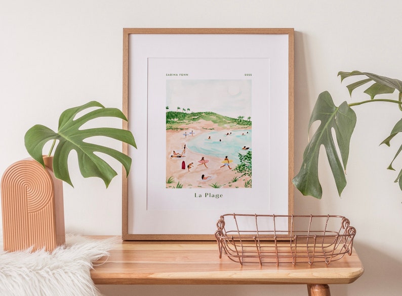 May include: A framed watercolor print of a beach scene with people swimming and sunbathing. The print is titled "La Plage" and is signed "Sabrina Ferr" and dated "2023".