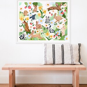 May include: A framed art print featuring a whimsical watercolor illustration of various rabbits amidst a floral and strawberry-filled garden. The artwork is in a white frame, and the overall color palette is soft and inviting.