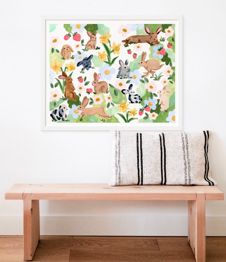 May include: A framed art print featuring a whimsical watercolor illustration of various rabbits amidst a floral and strawberry-filled garden. The artwork is in a white frame, and the overall color palette is soft and inviting.
