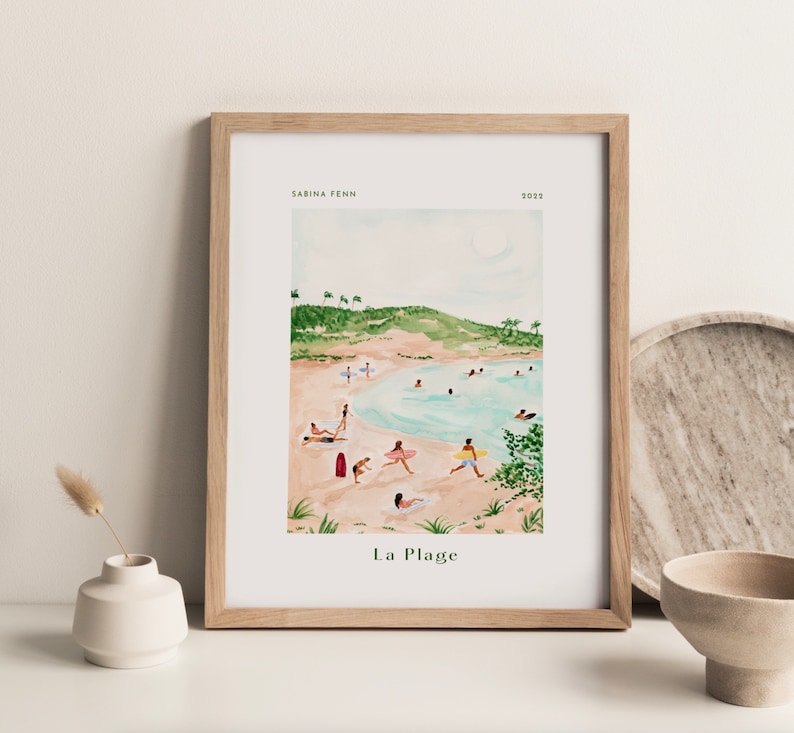 May include: A watercolor painting of a beach scene with people swimming and sunbathing. The painting is framed in a light wood frame and has the text "La Plage" at the bottom.