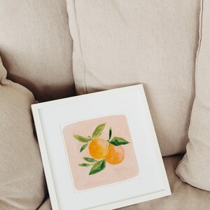 May include: A white framed painting of two orange citrus fruits with green leaves on a pink background.