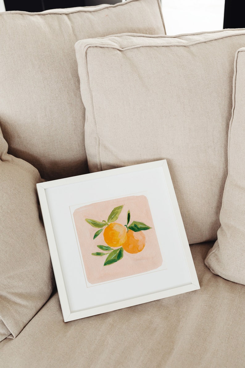 May include: A white framed painting of two orange citrus fruits with green leaves on a pink background.