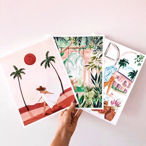 May include: Three watercolor prints featuring tropical scenes. The first print shows a woman in a white dress walking on a beach with palm trees and a red sun. The second print shows a lush green jungle with a stone archway. The third print shows a tropical house with palm trees and a blue sky.