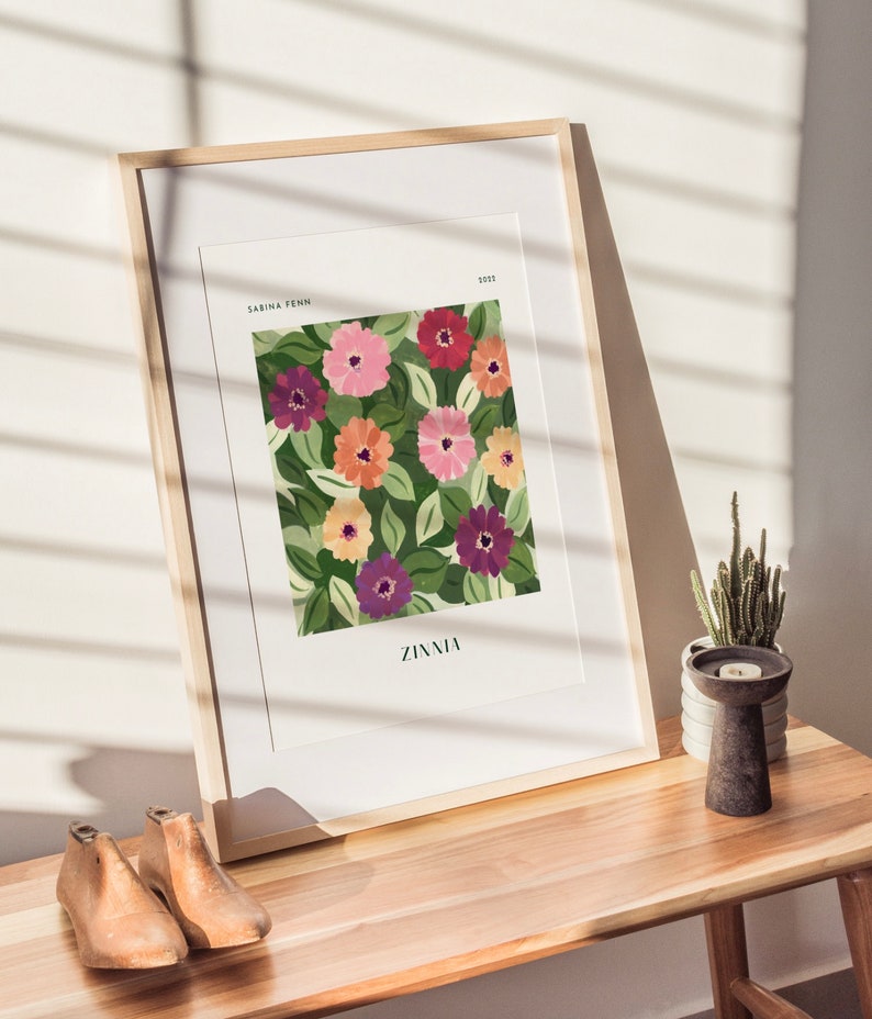 Puede incluir: Un cuadro enmarcado con un estampado floral de zinnias en tonos rosa, naranja y morado, con hojas verdes. La impresi&oacute;n se titula "Zinnia" y est&aacute; firmada "Sabina Fenn 2022".