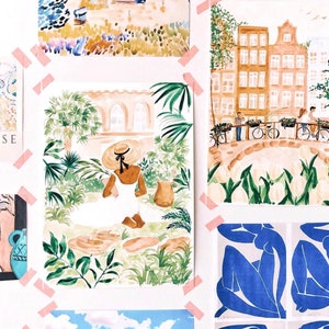 May include: A collection of watercolour art prints featuring a person in a white dress and straw hat sitting in a garden, a canal scene with buildings and bicycles, and blue abstract figures, all secured to a wall with strips of pink tape.