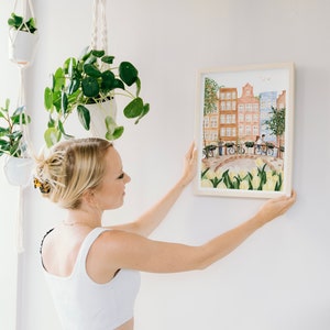 May include: A framed watercolour print depicting a canal scene with tall brown and blue buildings, a stone bridge, bicycles, and a foreground of yellow tulips. The artwork is enclosed in a light-coloured wooden frame with a clean, simple border.