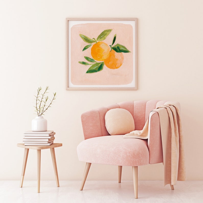 May include: A framed print of a painting of two orange citrus fruits with green leaves on a light pink background. The print is hanging on a white wall above a pink armchair with a throw blanket.