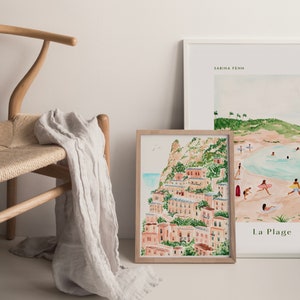 May include: Two framed watercolor paintings, one depicting a coastal scene with palm trees and the text "La Plage", the other depicting a hillside village with buildings and greenery.