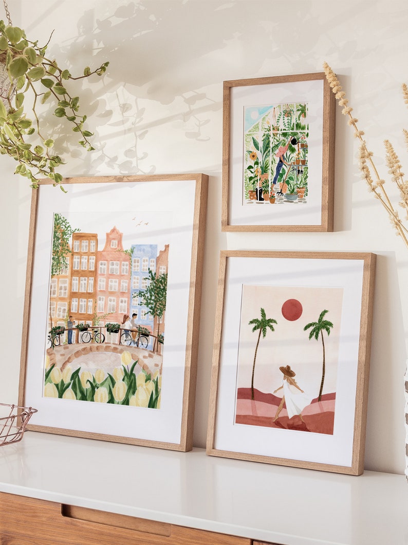 May include: Three framed art prints in light wood frames. The largest print depicts a canal scene with Dutch houses, bicycles, and yellow tulips. The top right print shows a person gardening in a greenhouse. The bottom right print features a person walking between palm trees under a red sun.