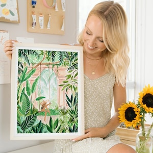 May include: A framed illustration of a woman sitting on a swing in a lush, tropical garden. The woman is wearing a straw hat and a white dress. The garden is filled with green plants and flowers. The frame is white and has a simple design.