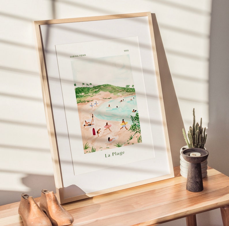 May include: A framed watercolor painting of a beach scene with people swimming and sunbathing. The painting is titled "La Plage" and is signed "Sabina Fenn 2023".