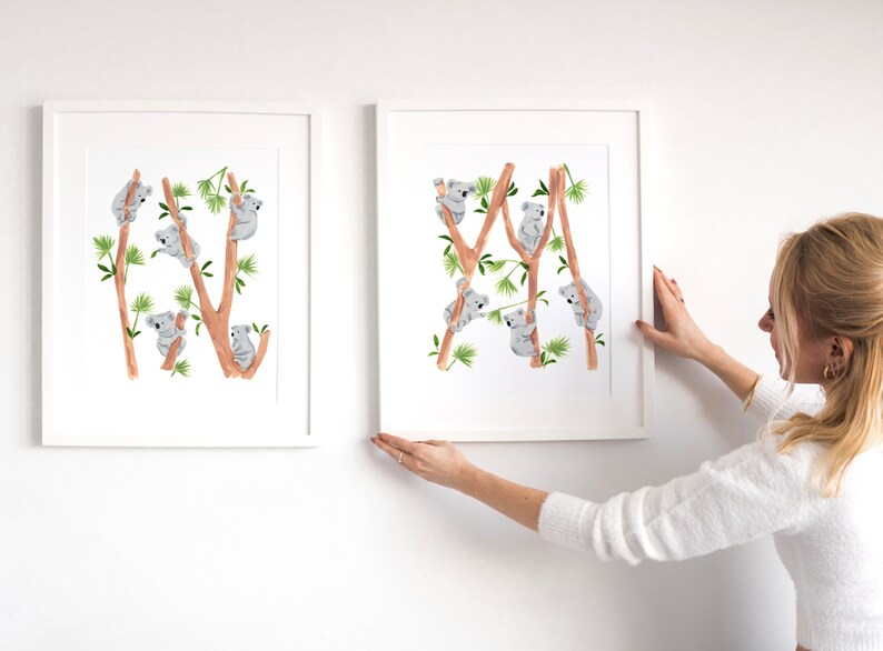 May include: Two framed prints of watercolor illustrations of koalas climbing on eucalyptus trees. The prints are white with a white mat and a white frame. The prints are hanging on a white wall.