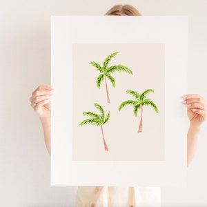 May include: A framed art print featuring three watercolor palm trees. The palm trees have green fronds and brown trunks, set against a light beige background. The artwork is displayed in a white mat and frame, suitable for tropical decor.