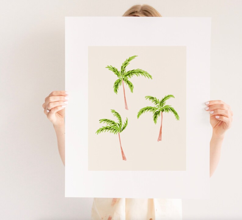 May include: A framed art print featuring three watercolor palm trees. The palm trees have green fronds and brown trunks, set against a light beige background. The artwork is displayed in a white mat and frame, suitable for tropical decor.