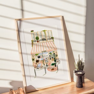 Op de afbeelding: Een ingelijst aquarel schilderij van een Parijse cafe met een roze en wit gestreept luifel. De cafe heeft een bord met de tekst "Cafe" en "Patisserie". Er zitten twee mensen aan een tafeltje buiten de cafe.