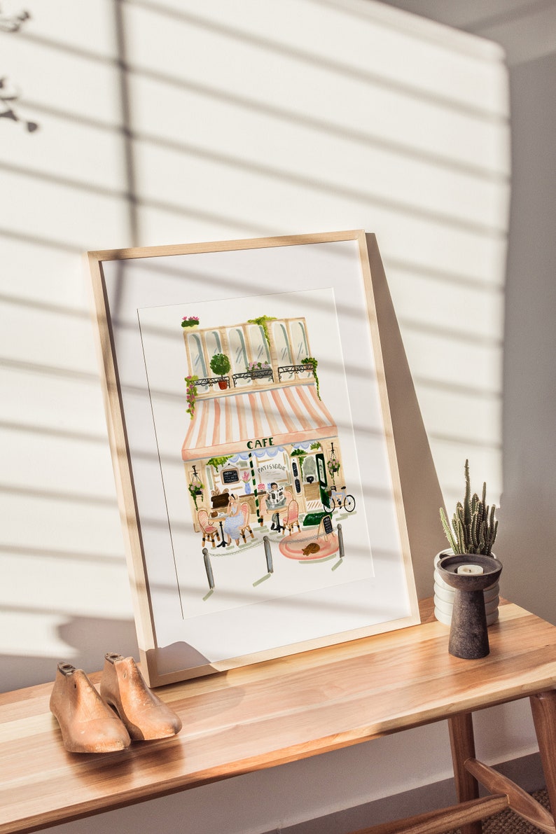 Op de afbeelding: Een ingelijst aquarel schilderij van een Parijse cafe met een roze en wit gestreept luifel. De cafe heeft een bord met de tekst "Cafe" en "Patisserie". Er zitten twee mensen aan een tafeltje buiten de cafe.
