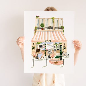 Op de afbeelding: Een aquarel-illustratie van een Parijse cafe met een gestreept luifel, een bord met de tekst "Cafe Patisserie", en twee mensen die aan tafels buiten zitten. Er staat een fiets voor het cafe en een kat zit op een roze kleed.
