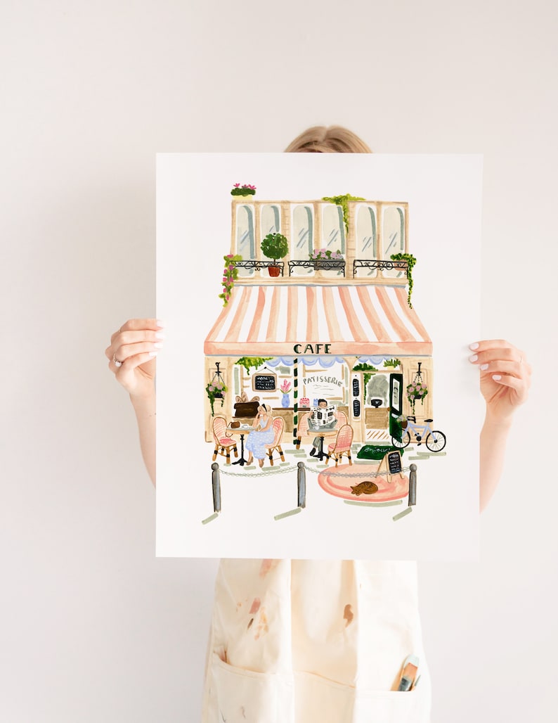 Op de afbeelding: Een aquarel-illustratie van een Parijse cafe met een gestreept luifel, een bord met de tekst "Cafe Patisserie", en twee mensen die aan tafels buiten zitten. Er staat een fiets voor het cafe en een kat zit op een roze kleed.