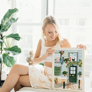 K&ouml;nnte beinhalten: Eine Frau h&auml;lt eine gerahmte Illustration eines bunten Geb&auml;udes mit einer gr&uuml;nen T&uuml;r, Topfpflanzen und einer schwarzen Katze auf einem Fenstersims. Das Geb&auml;ude hat eine gekachelte Fassade und einen wei&szlig;en Rahmen.