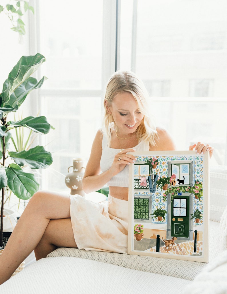 K&ouml;nnte beinhalten: Eine Frau h&auml;lt eine gerahmte Illustration eines bunten Geb&auml;udes mit einer gr&uuml;nen T&uuml;r, Topfpflanzen und einer schwarzen Katze auf einem Fenstersims. Das Geb&auml;ude hat eine gekachelte Fassade und einen wei&szlig;en Rahmen.