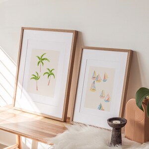 May include: Two framed art prints. One features three green palm trees with pink trunks on a cream background. The other print shows colorful sailboats on a cream background. Both are in light brown wooden frames.