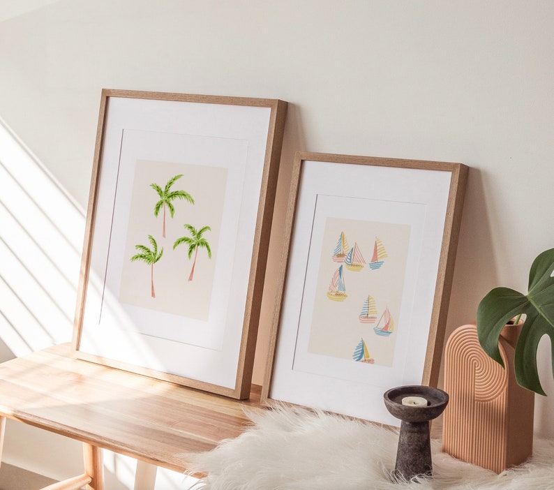 May include: Two framed art prints. One features three green palm trees with pink trunks on a cream background. The other print shows colorful sailboats on a cream background. Both are in light brown wooden frames.