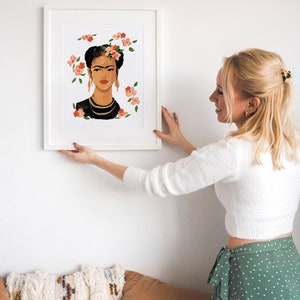 May include: A white framed print of a painting of Frida Kahlo with flowers in her hair. The painting is on a white wall and a person is hanging it up.